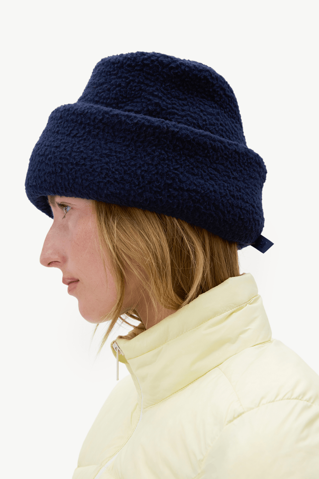 A side-profile view of a woman wearing a dark navy blue fuzzy fleece beanie and a pale yellow puffer jacket against a plain white background.