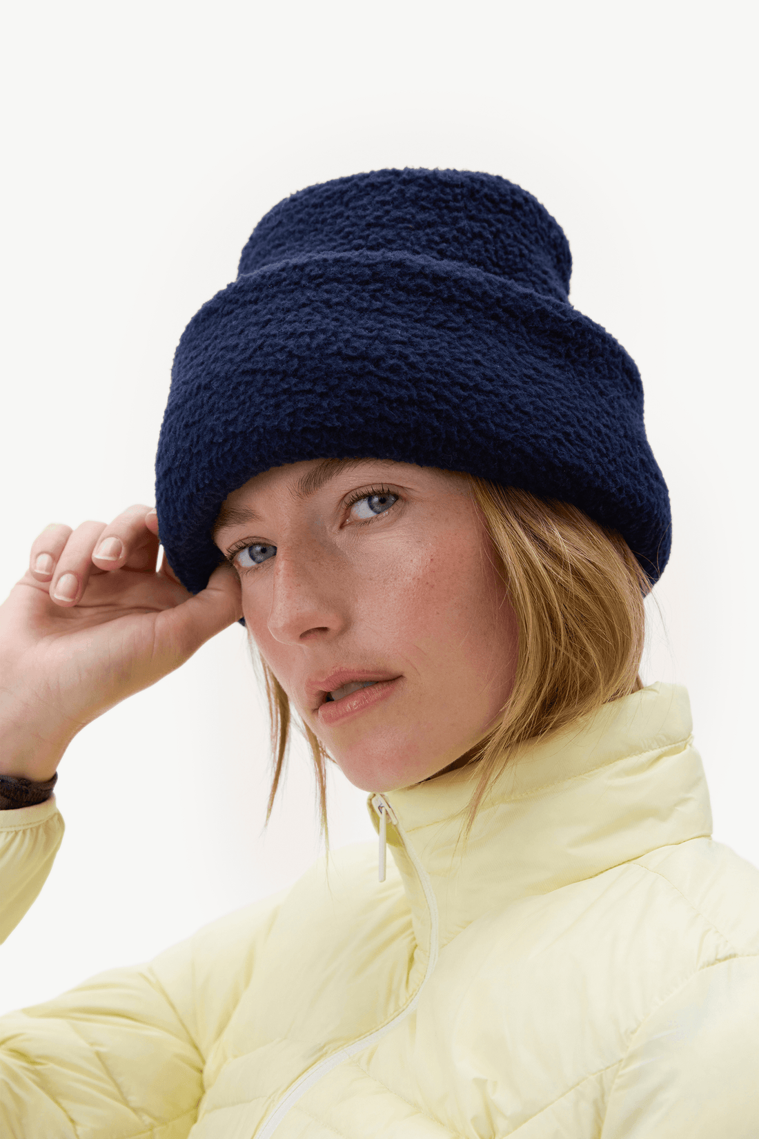 A woman wearing a dark navy blue bouclé beanie and a pale yellow puffer jacket, posing against a clean white background.