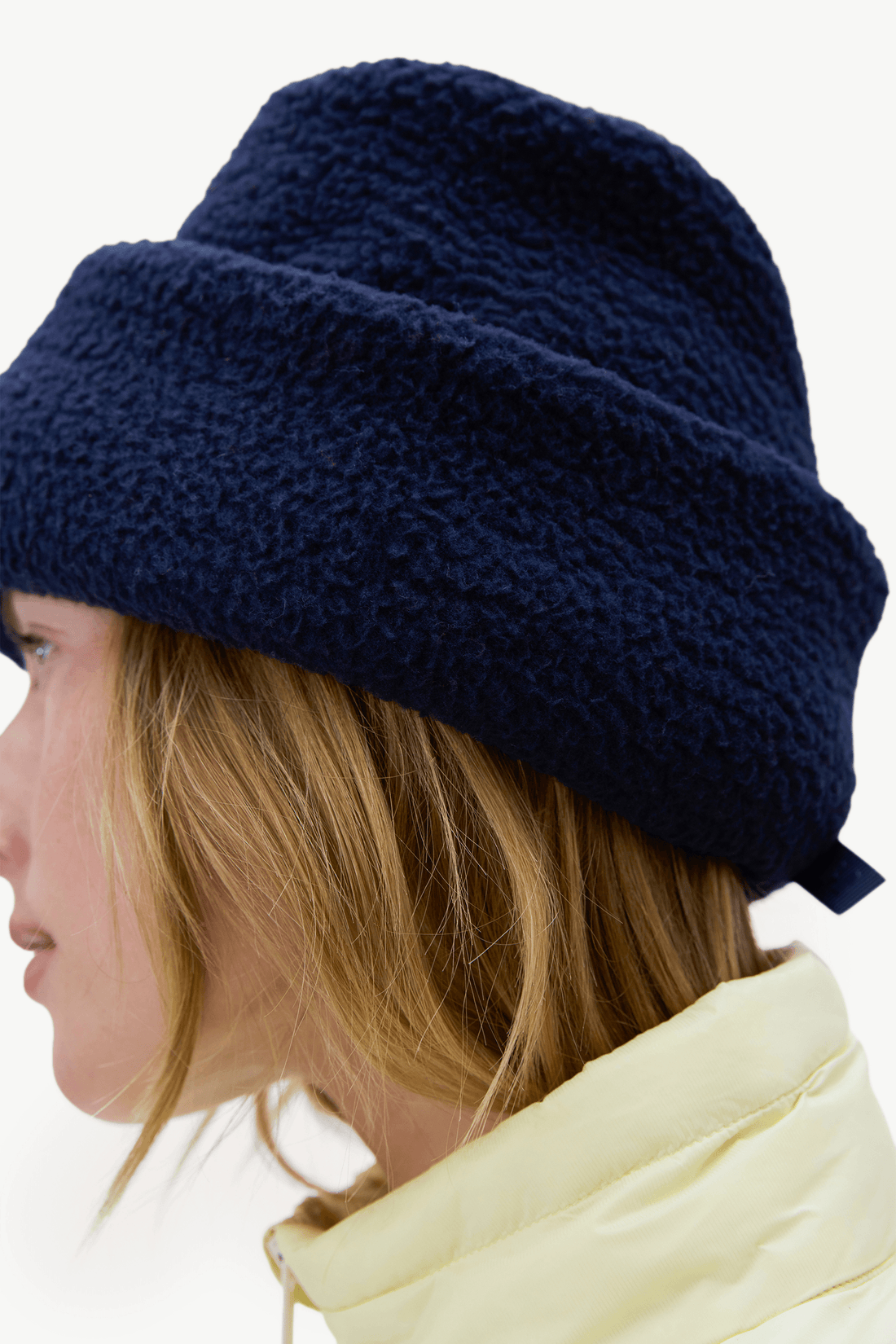 A close-up side profile of a person wearing a dark navy blue textured fleece beanie and a pale yellow puffer jacket against a plain white background.