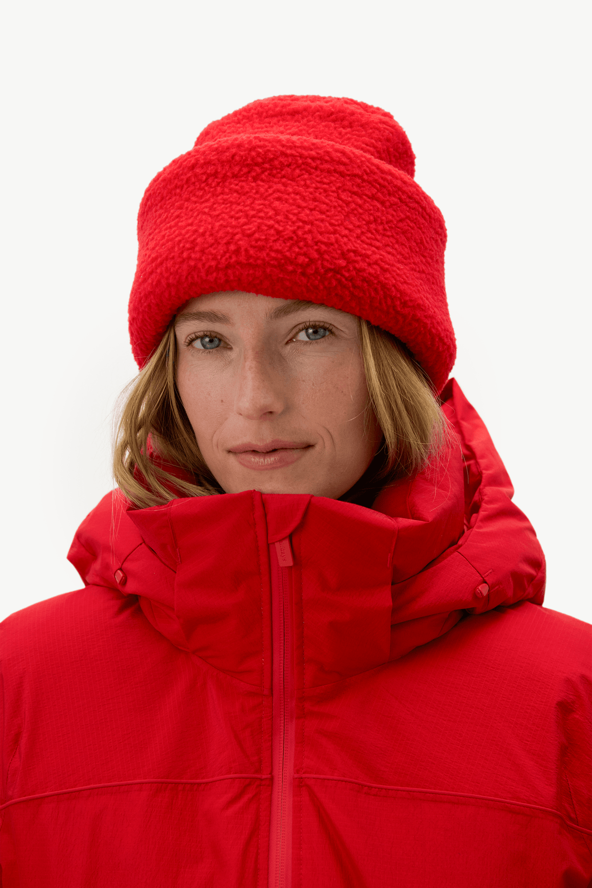 A close-up of a model wearing a bright red fleece beanie and a matching red puffer jacket against a clean white background.