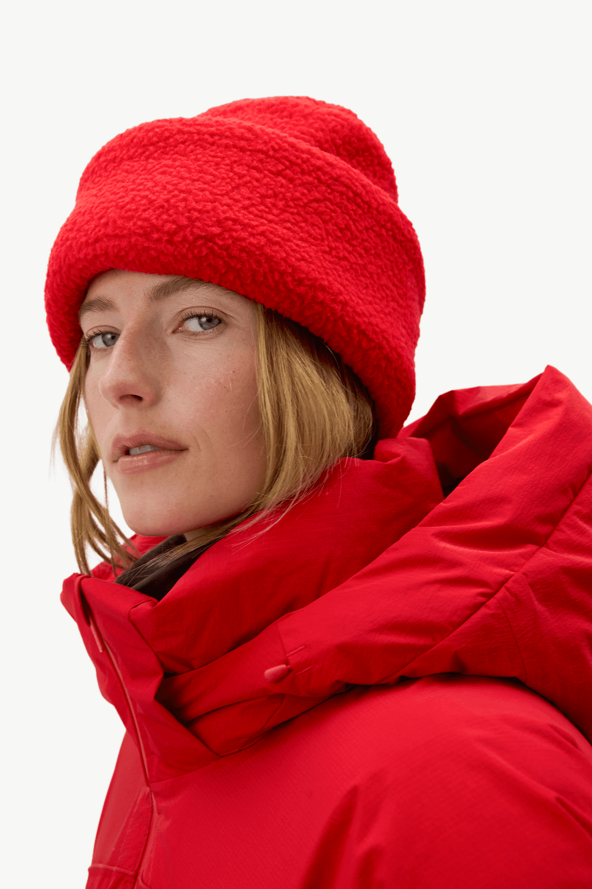 A close-up of a woman wearing a bright red fuzzy beanie and a matching red hooded jacket against a clean white background.
