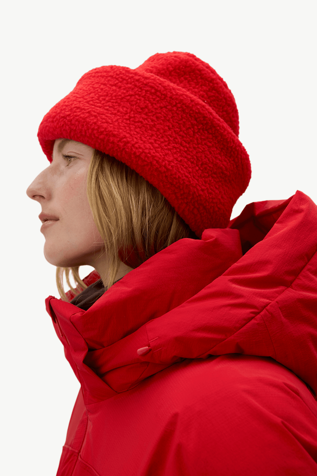 A close-up side profile of a person wearing a bright red fuzzy knit beanie and a matching red hooded winter jacket against a plain light background.