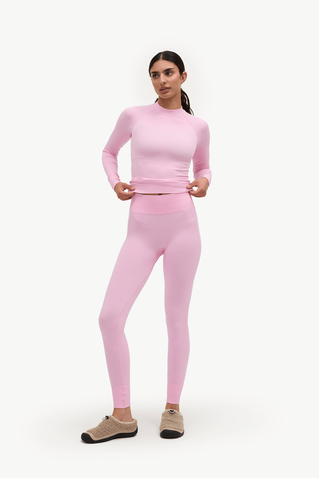 A woman modeling a matching light pink two-piece activewear set consisting of a long-sleeve mock-neck top and high-waisted leggings, paired with beige slip-on shoes, against a plain white studio background.