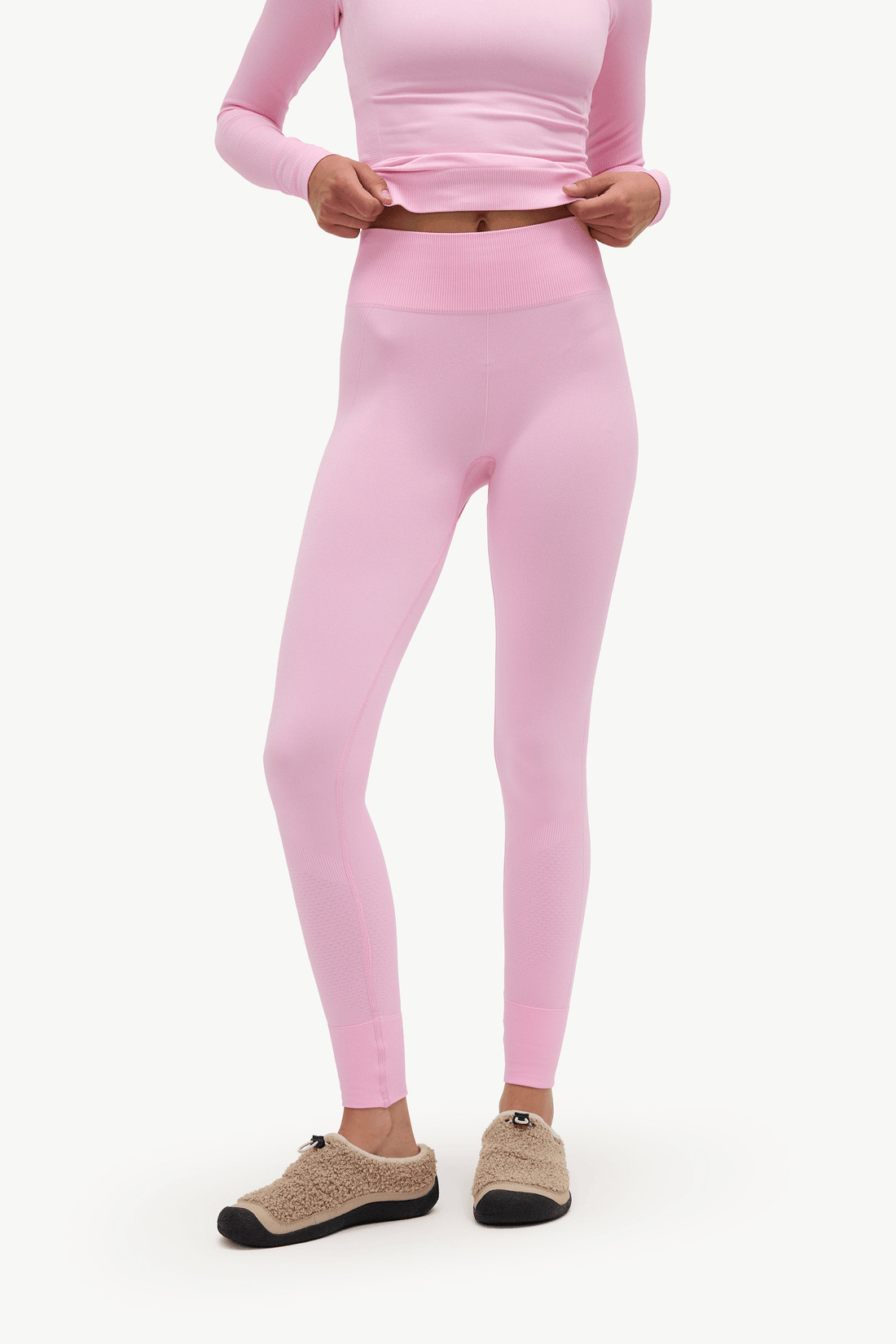 A model wearing light pink high-waisted leggings and a matching long-sleeve top, paired with beige knit slip-on shoes, against a plain white background.