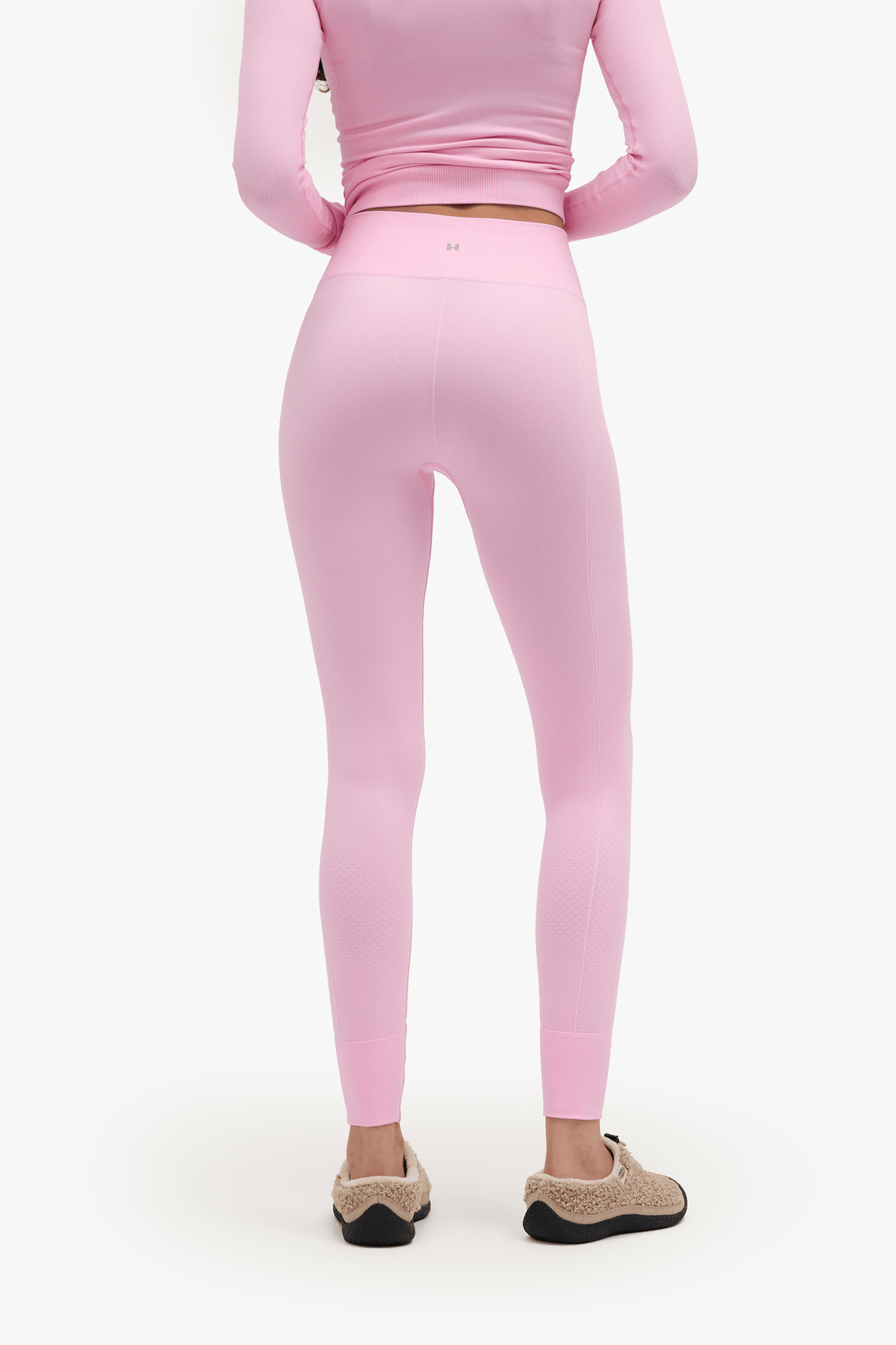 A model shown from the back wearing a matching light pink long-sleeve crop top and high-waisted leggings, paired with beige slip-on slippers, against a plain white background.