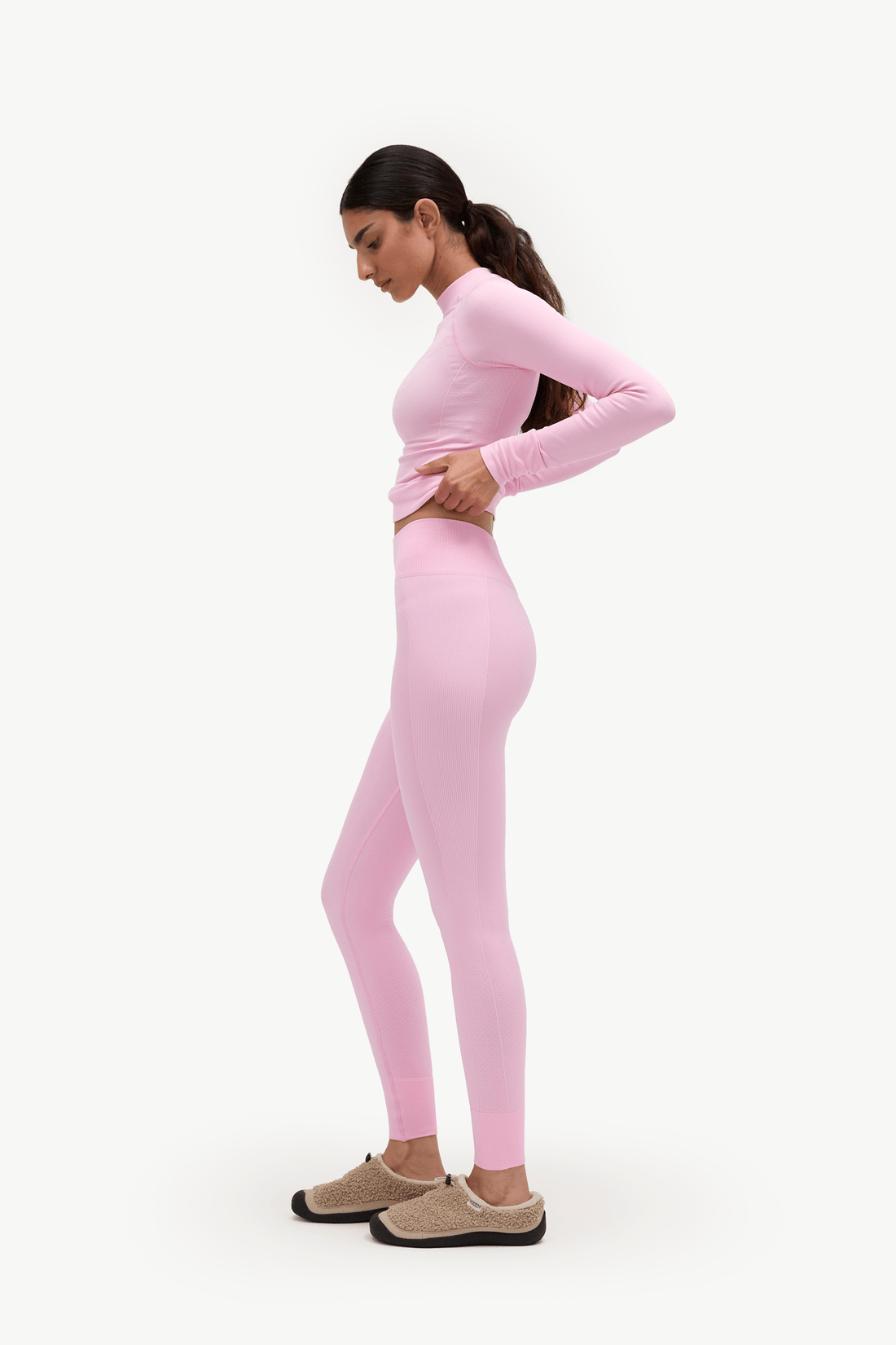 A woman modeling a light pink long-sleeve mock-neck top and matching high-waisted leggings, paired with beige slip-on shoes, shown in profile against a plain white background.