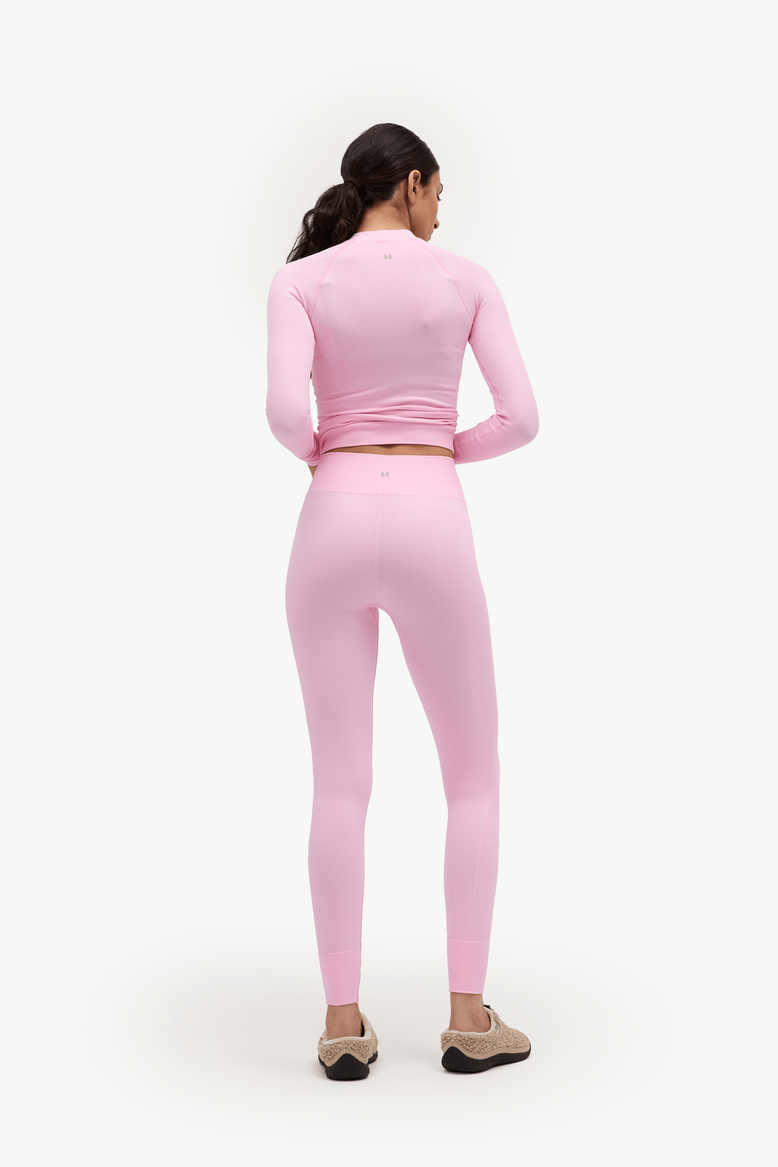 A woman is shown from the back wearing a matching light pink two-piece activewear set consisting of a long-sleeve crop top and high-waisted leggings, paired with beige slip-on shoes against a plain white studio background.
