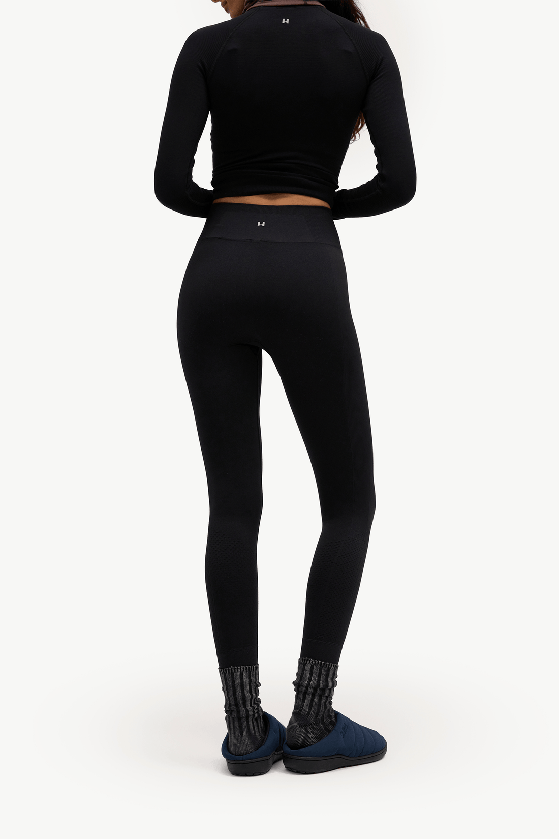 Rear view of a model wearing a black long-sleeve cropped athletic top and black high-waisted leggings, paired with dark blue slip-on shoes and textured black socks, against a plain white studio background.