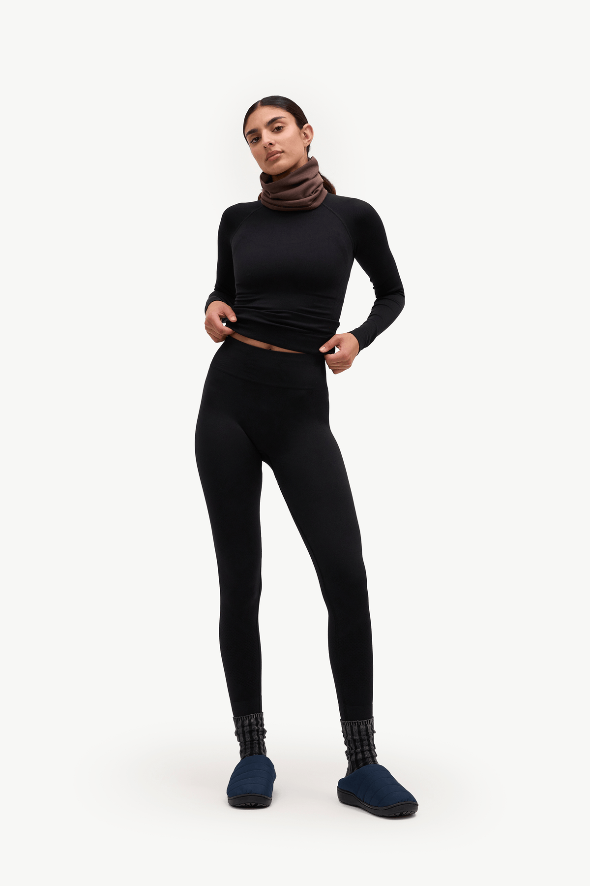 A woman modeling a black long-sleeve turtleneck top and matching black leggings, styled with a brown cowl neck warmer, dark blue slippers, and patterned socks, against a plain white studio background.