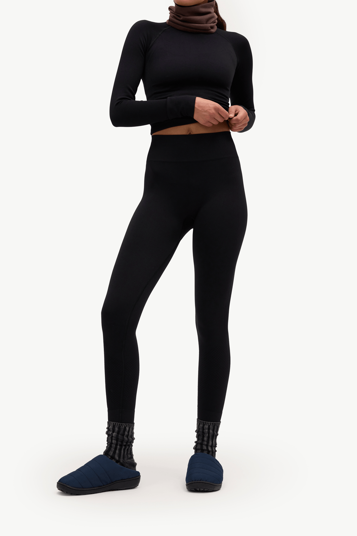 A model wearing a black long-sleeve crop top, black high-waisted leggings, dark blue slip-on slippers, and a brown neck gaiter, standing against a plain white studio background.