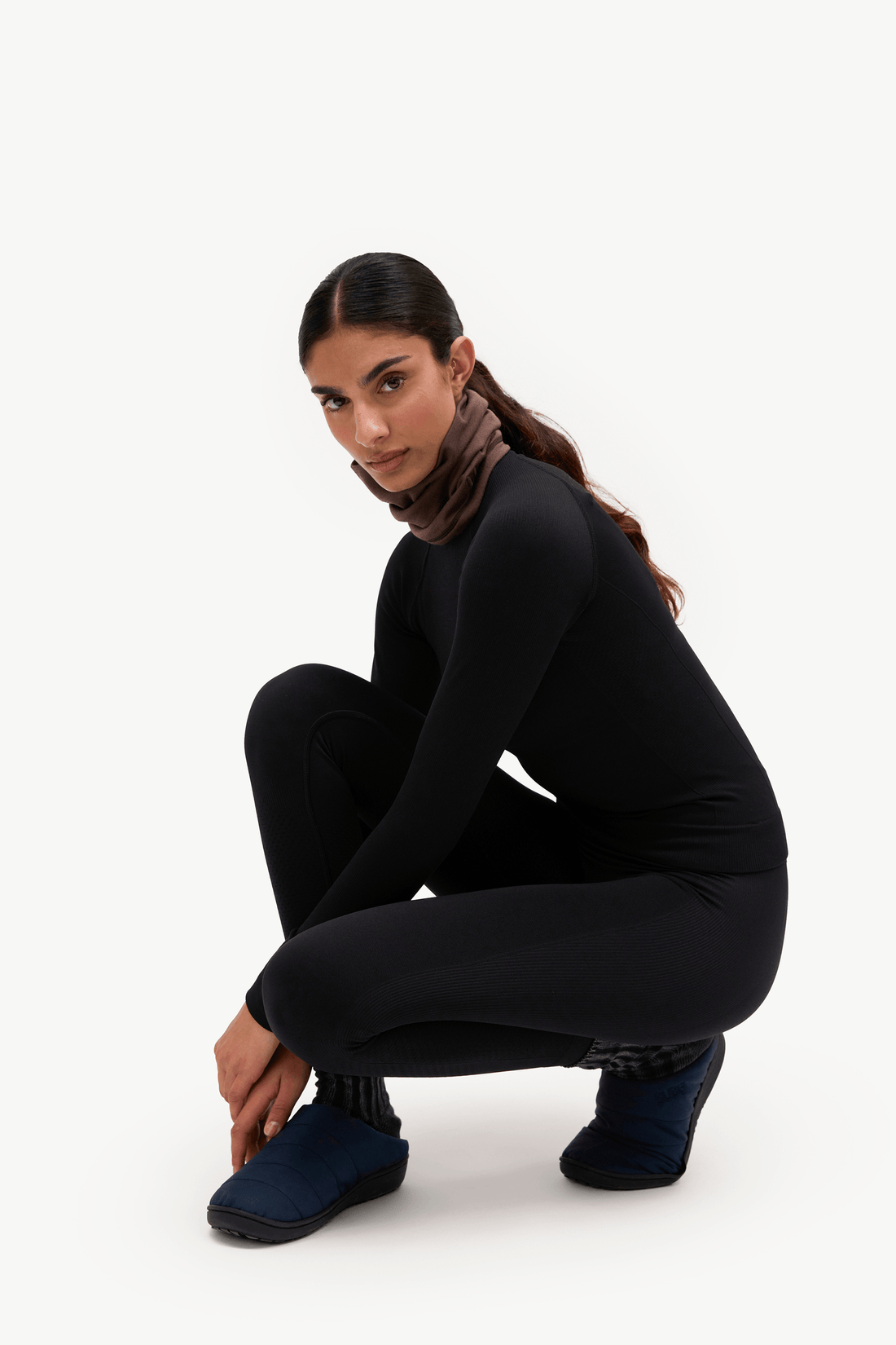 A woman crouching in a black long-sleeve top, black leggings, and dark blue slip-on shoes against a plain white studio background.