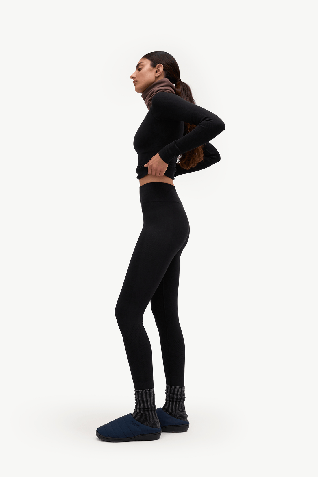 A woman in a black long-sleeve crop top, black leggings, and dark blue slippers stands in profile against a plain white studio background.