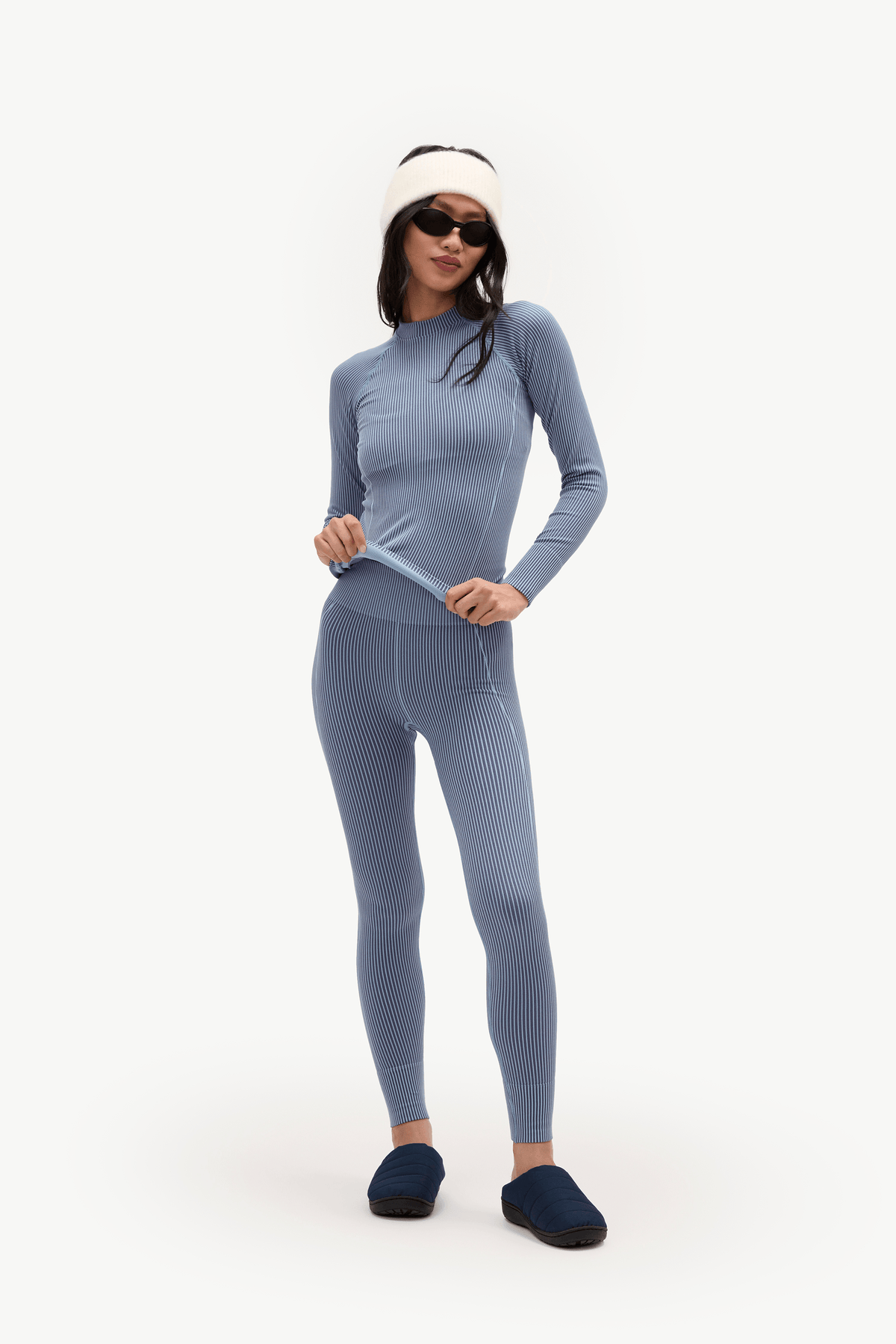 A woman modeling a matching ribbed knit two-piece outfit in dusty blue, consisting of a long-sleeve top and leggings, styled with a cream beanie, black sunglasses, and dark blue slippers against a plain white studio background.