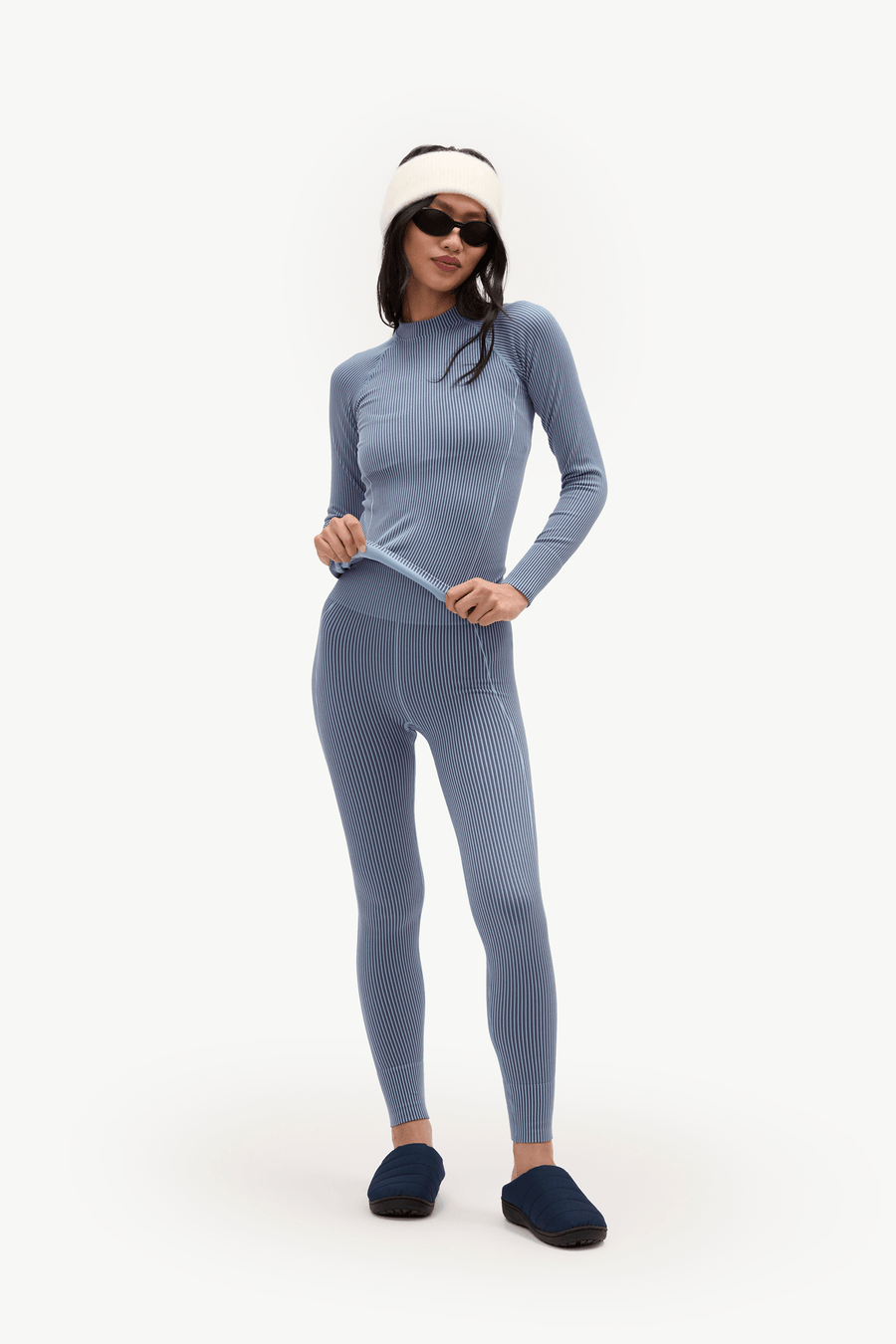 A woman modeling a matching ribbed knit two-piece outfit in dusty blue, consisting of a long-sleeve top and leggings, styled with a cream beanie, black sunglasses, and dark blue slippers against a plain white studio background.