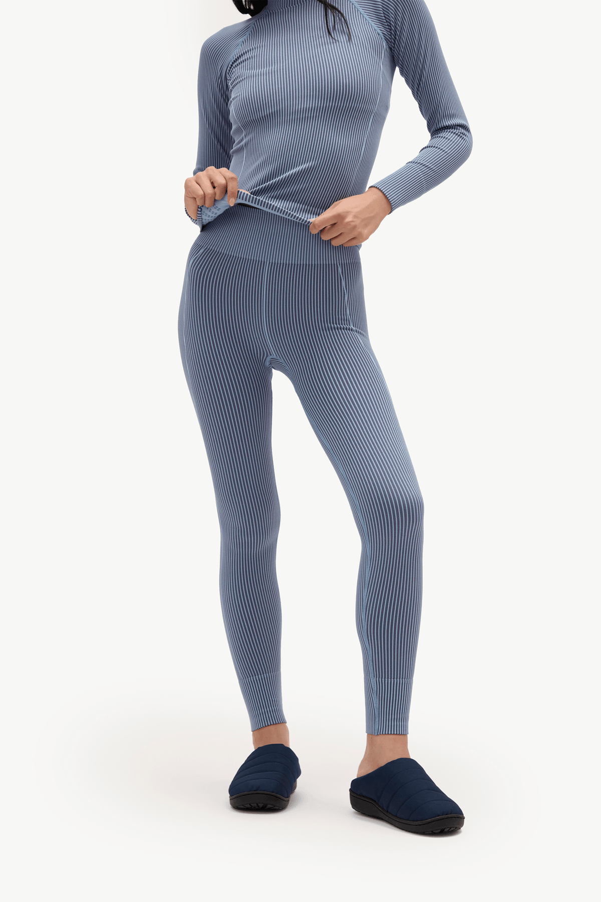 A model wearing a matching ribbed long-sleeve top and leggings set in a dusty slate blue color, paired with dark blue slip-on slippers, against a plain white studio background.