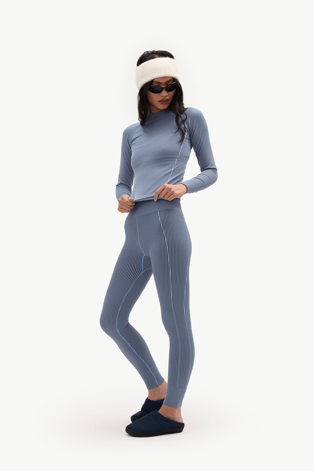 A woman modeling a matching ribbed knit one-piece outfit in a dusty blue-gray color, featuring a long-sleeve mock neck top and high-waisted leggings with light piping details. She is also wearing a cream headband, black sunglasses, and dark blue slippers against a plain white studio background.