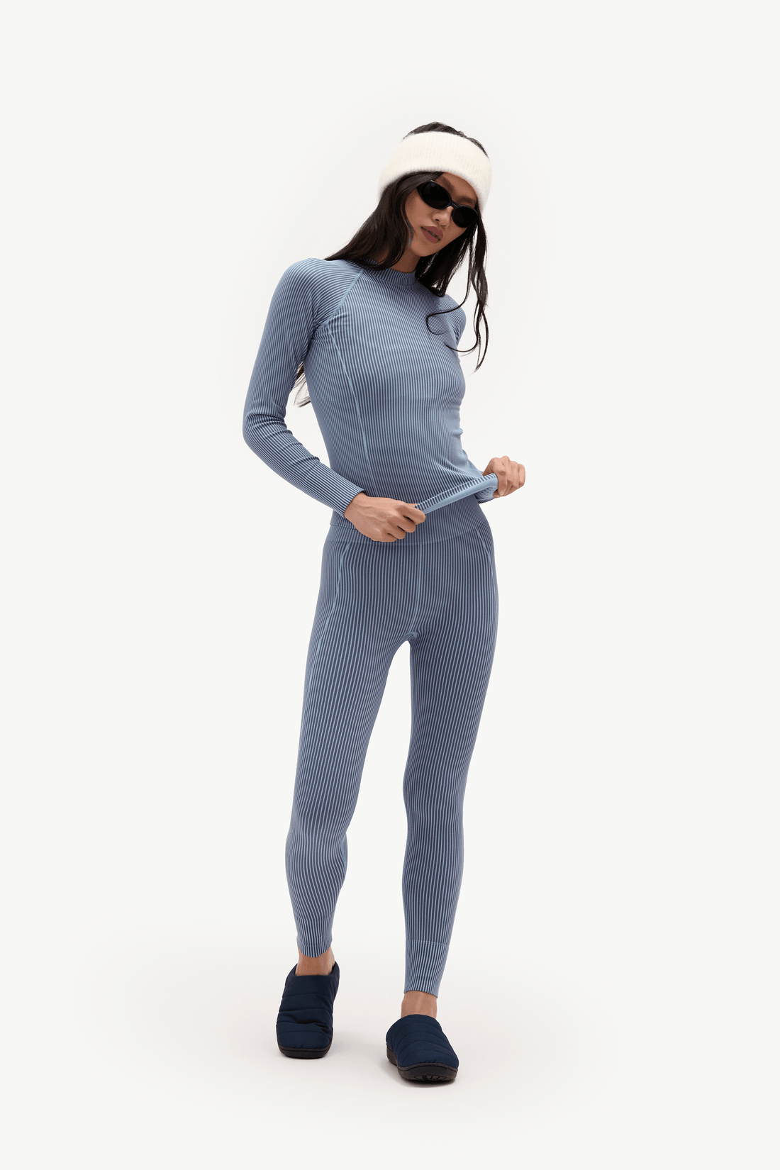 A woman modeling a light blue ribbed one-piece jumpsuit or wetsuit-style full-body suit, paired with a cream beanie, dark sunglasses, and dark slip-on shoes, against a plain white studio background.