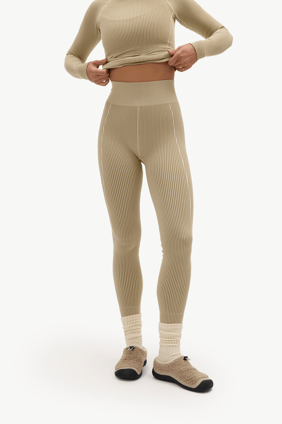 A model wearing beige ribbed high-waisted leggings and a matching long-sleeve crop top, paired with cream socks and beige slip-on shoes, against a plain white studio background.