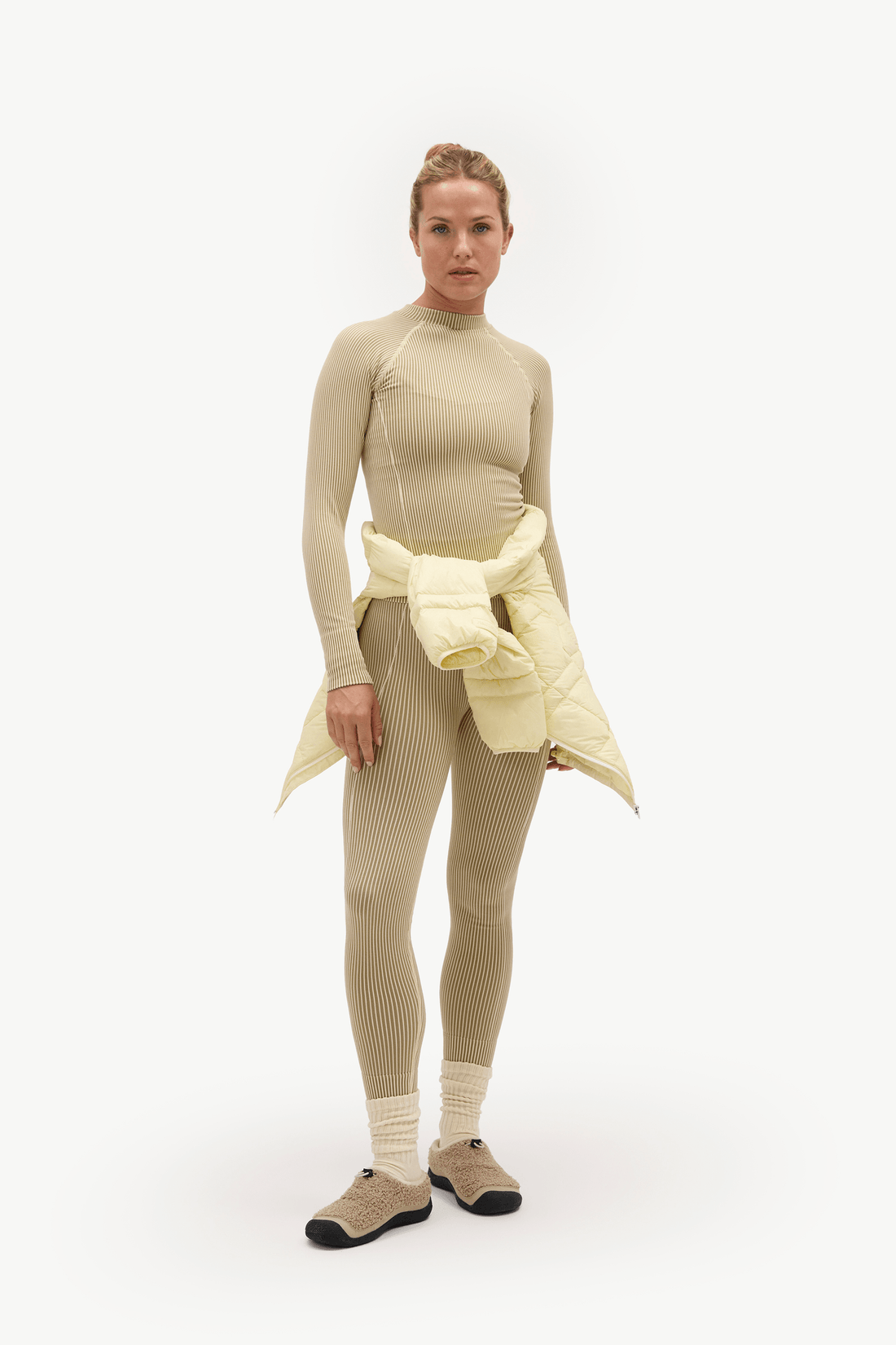 A model stands against a plain white studio background wearing a fitted beige long-sleeve top and matching ribbed leggings, with a pale yellow puffer jacket tied around her waist and beige socks with dark shoes.