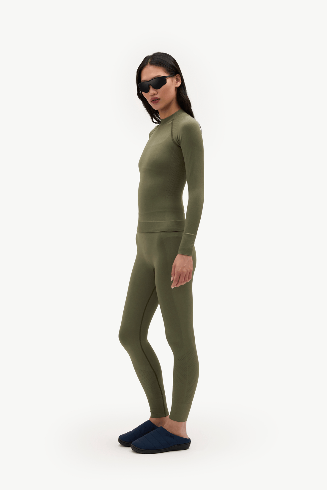 A woman modeling a matching olive green long-sleeve top and leggings set, paired with dark blue slippers and black sunglasses, against a plain white background.