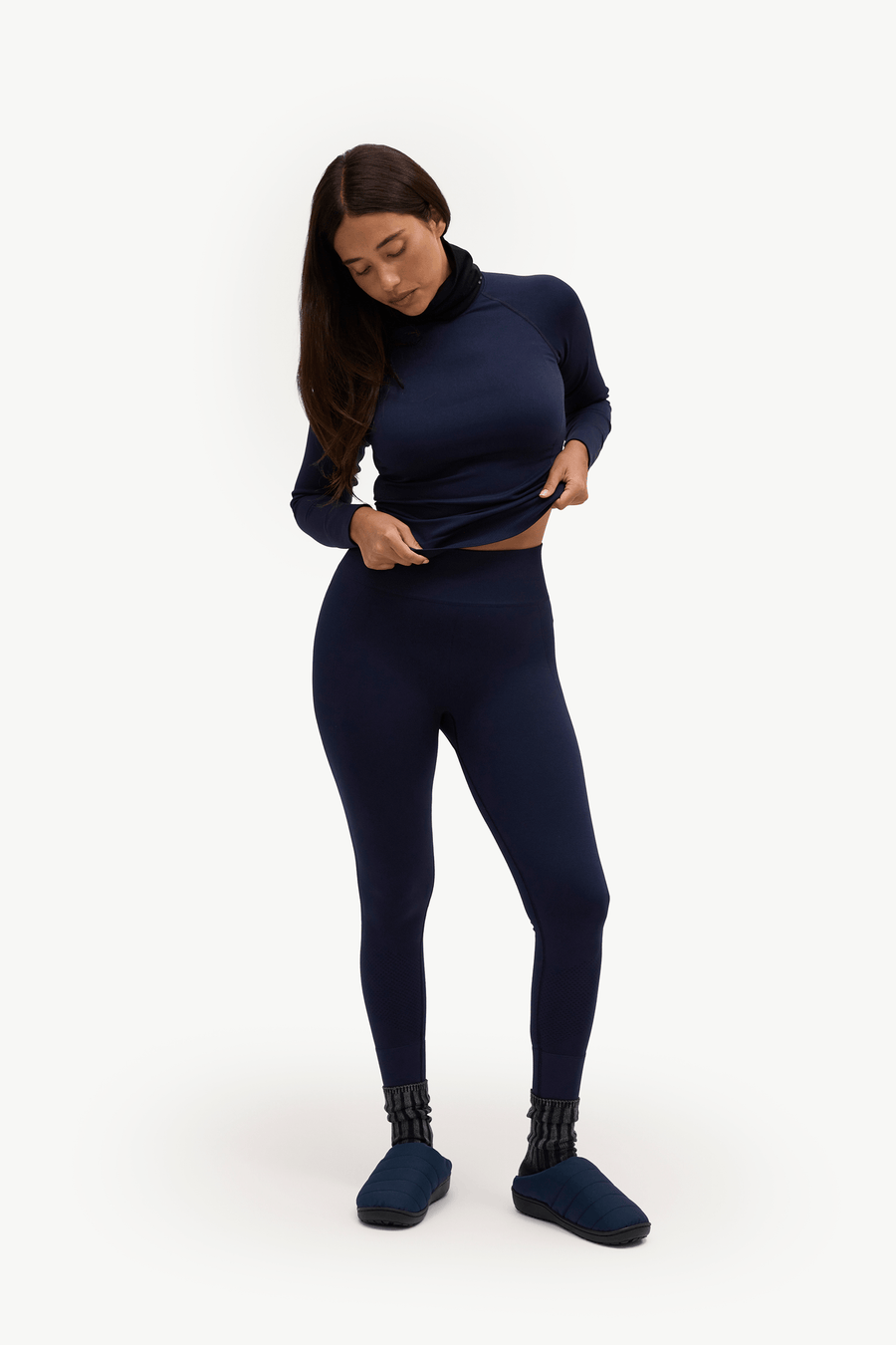 A woman modeling a matching navy blue activewear set consisting of a long-sleeve mock neck top and high-waisted leggings, paired with matching navy socks, against a plain white studio background.