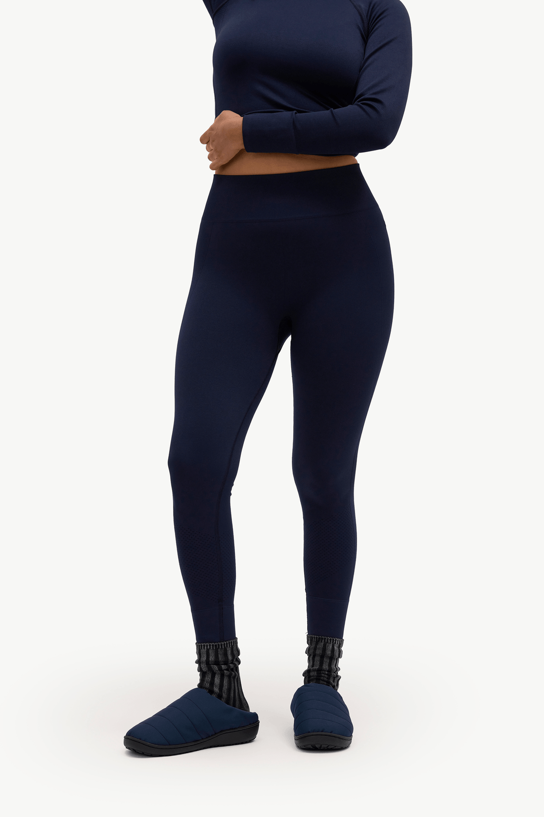 A model wearing navy blue high-waisted leggings, a matching long-sleeve crop top, and navy slip-on slippers, shown from the waist down against a plain white studio background.