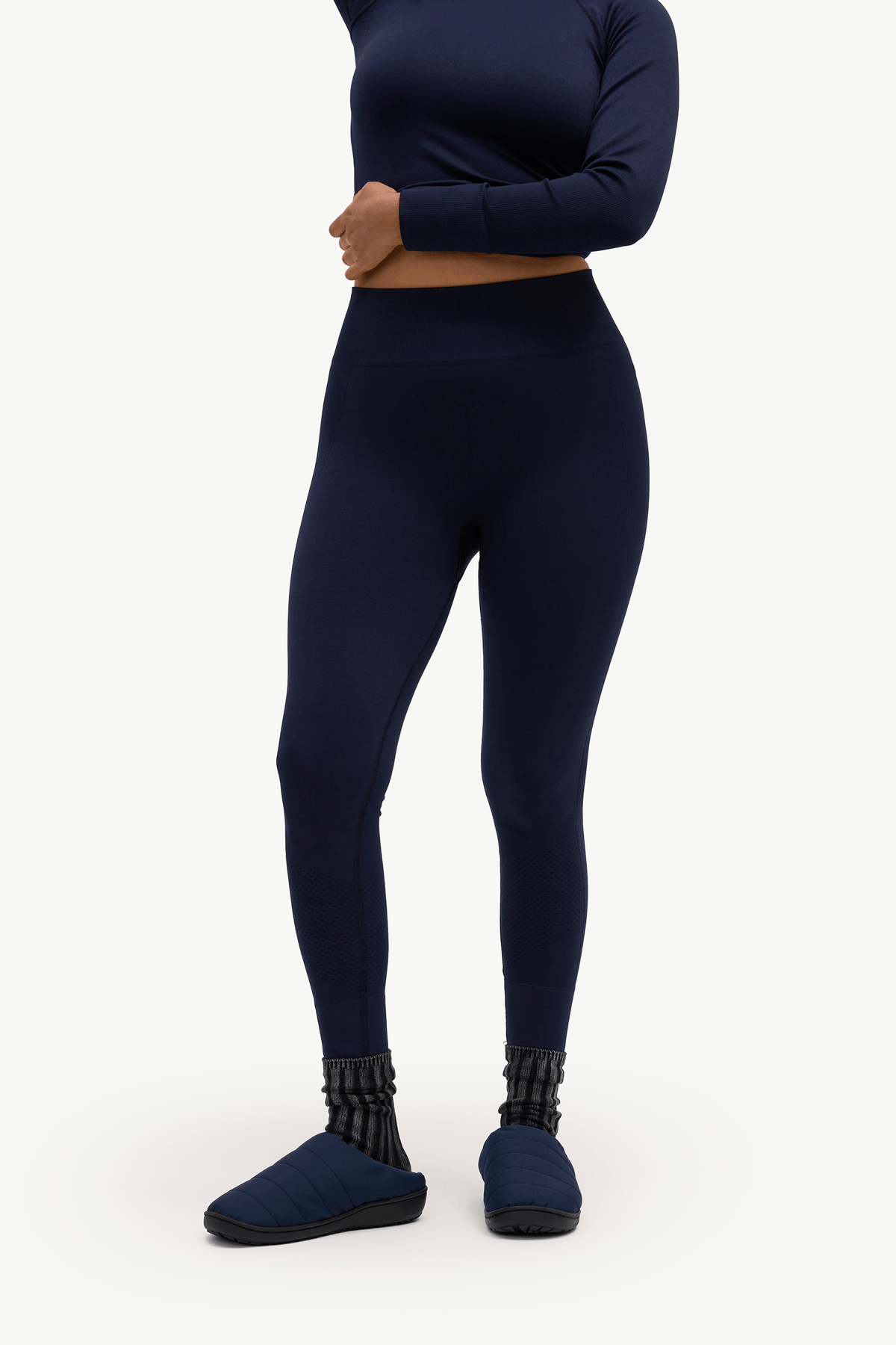 A model wearing navy blue high-waisted leggings, a matching long-sleeve crop top, and navy slip-on slippers, shown from the waist down against a plain white studio background.
