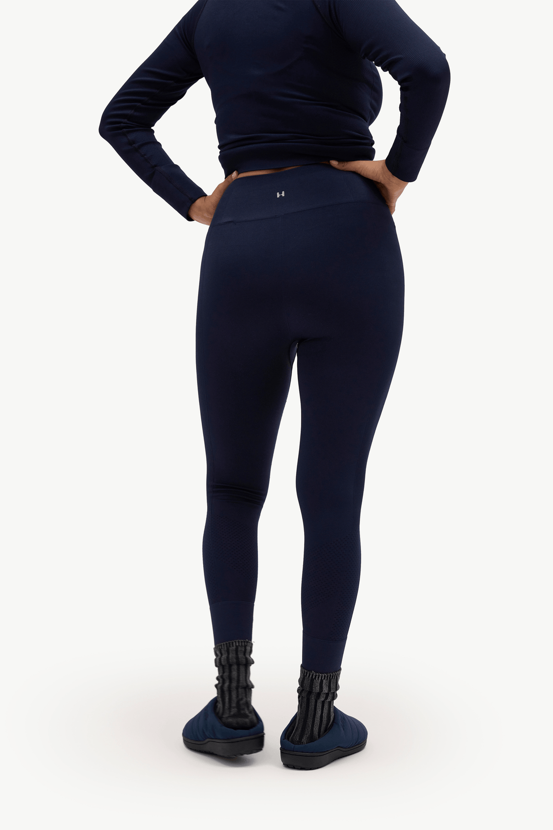 A model from the back wearing navy blue leggings, a matching navy long-sleeve top, and dark slip-on shoes against a plain white studio background.