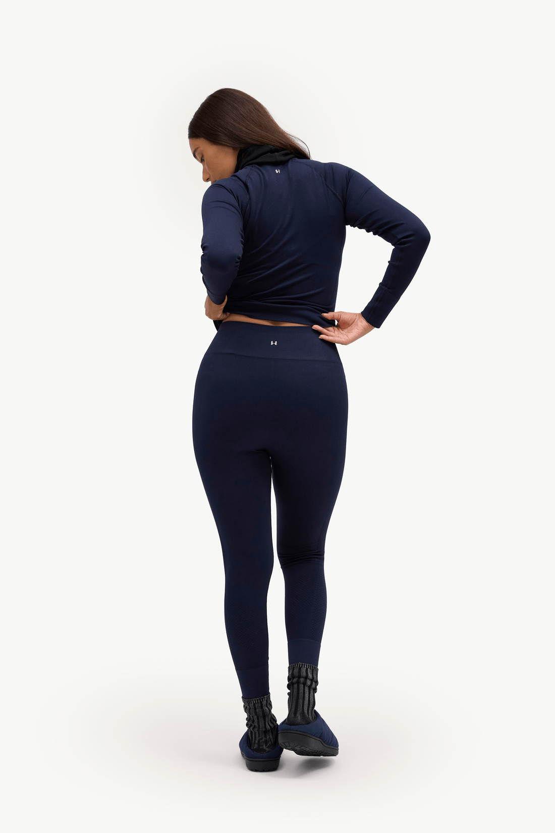 A woman is shown from behind wearing a matching navy blue activewear set consisting of a long-sleeve cropped top and high-waisted leggings, paired with black platform boots, against a plain white studio background.