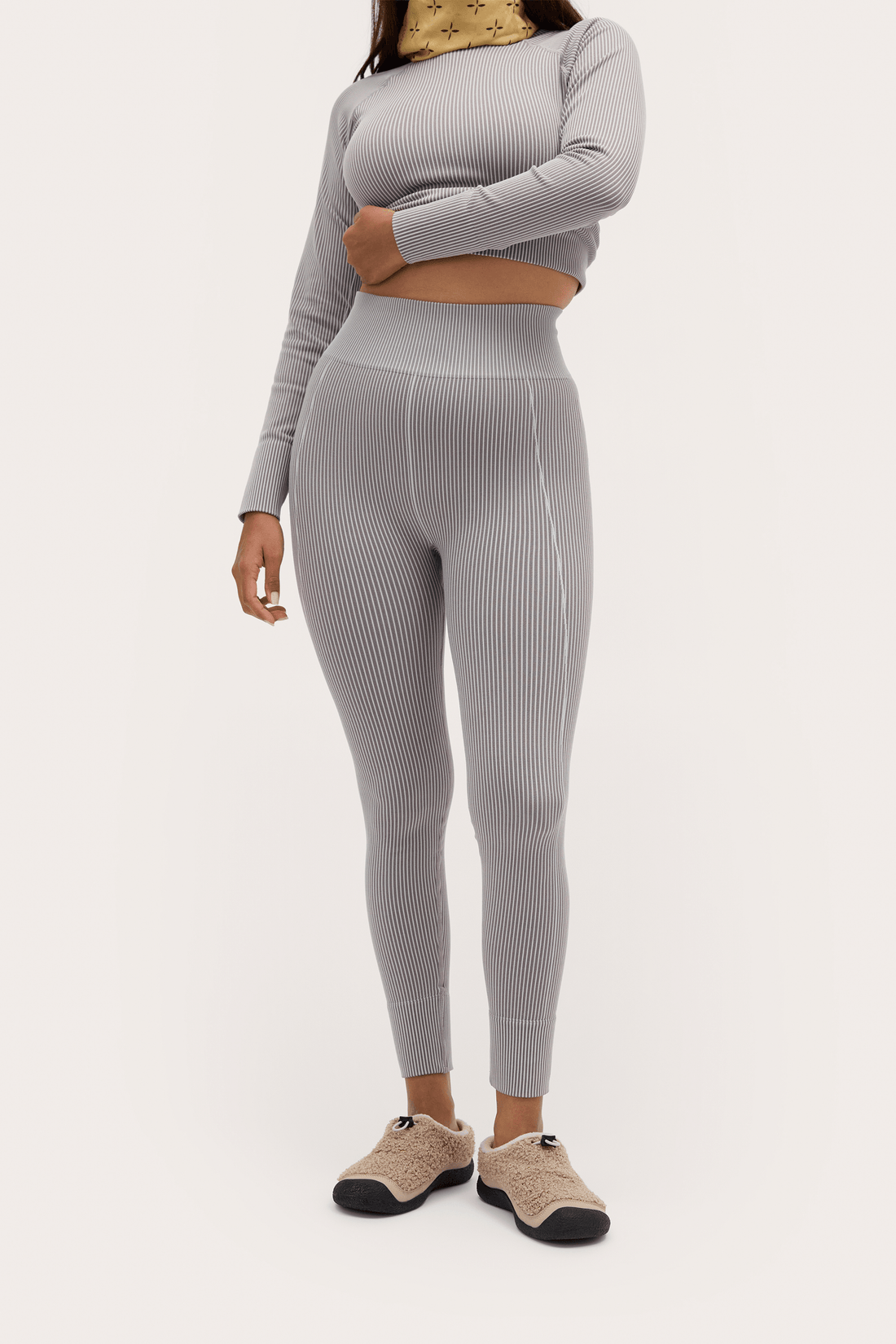A model wearing a light gray ribbed two-piece activewear set consisting of a long-sleeve crop top and high-waisted leggings, paired with beige slip-on shoes, against a plain off-white background.