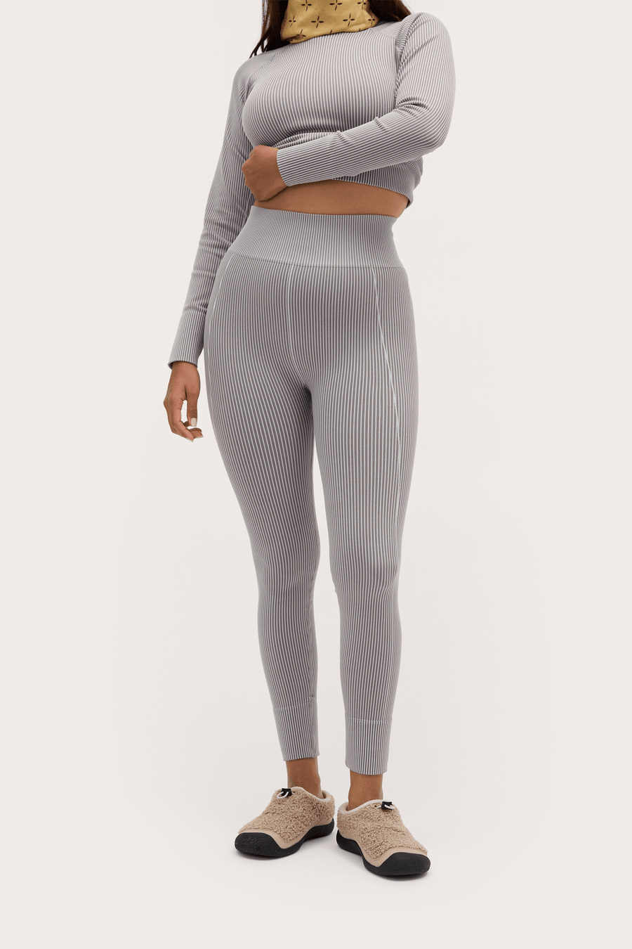 A model wearing a light gray ribbed two-piece activewear set consisting of a long-sleeve crop top and high-waisted leggings, paired with beige slip-on shoes, against a plain off-white background.