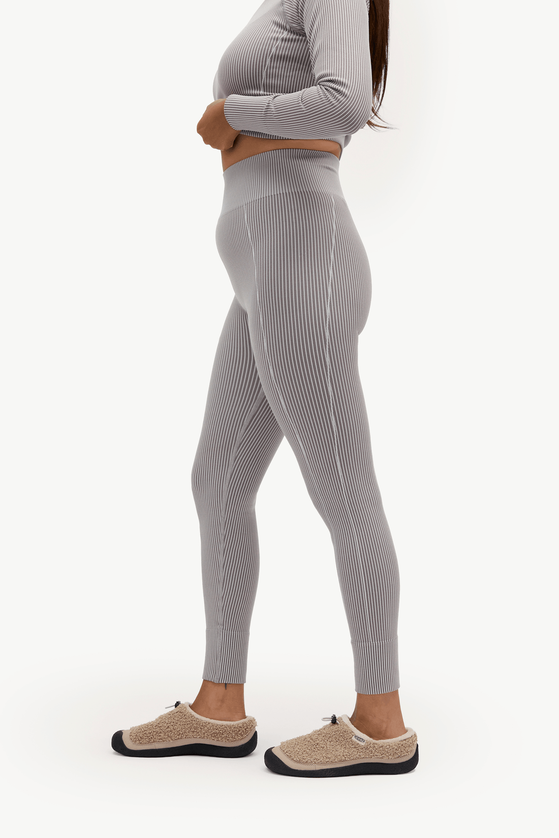 A side-profile view of a model wearing high-waisted, ribbed gray leggings, a matching gray long-sleeve crop top, and beige slip-on slippers against a plain white background.