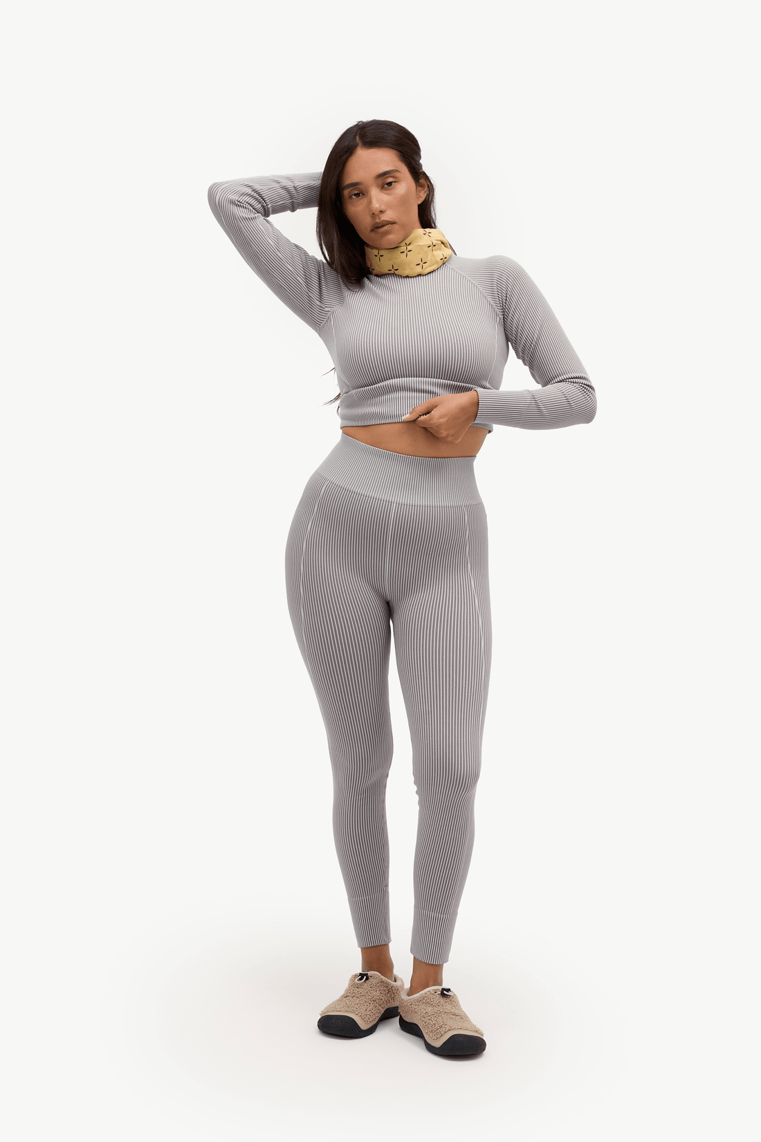 A woman modeling a light gray ribbed activewear set consisting of a long-sleeve crop top and matching high-waisted leggings, paired with beige sneakers, against a plain white studio background.