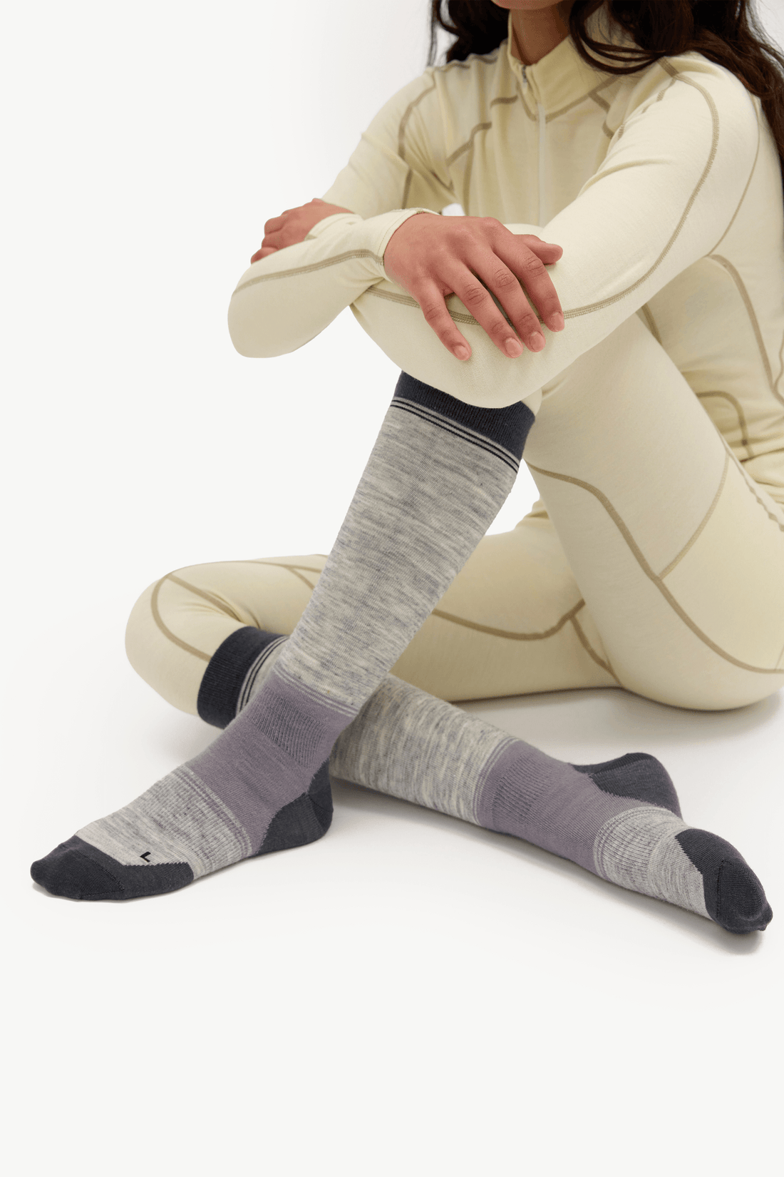 A model wearing a cream-colored athletic zip-up jacket and matching leggings sits cross-legged on a white studio floor, showcasing a pair of heather gray knee-high socks with darker gray and lavender sections.