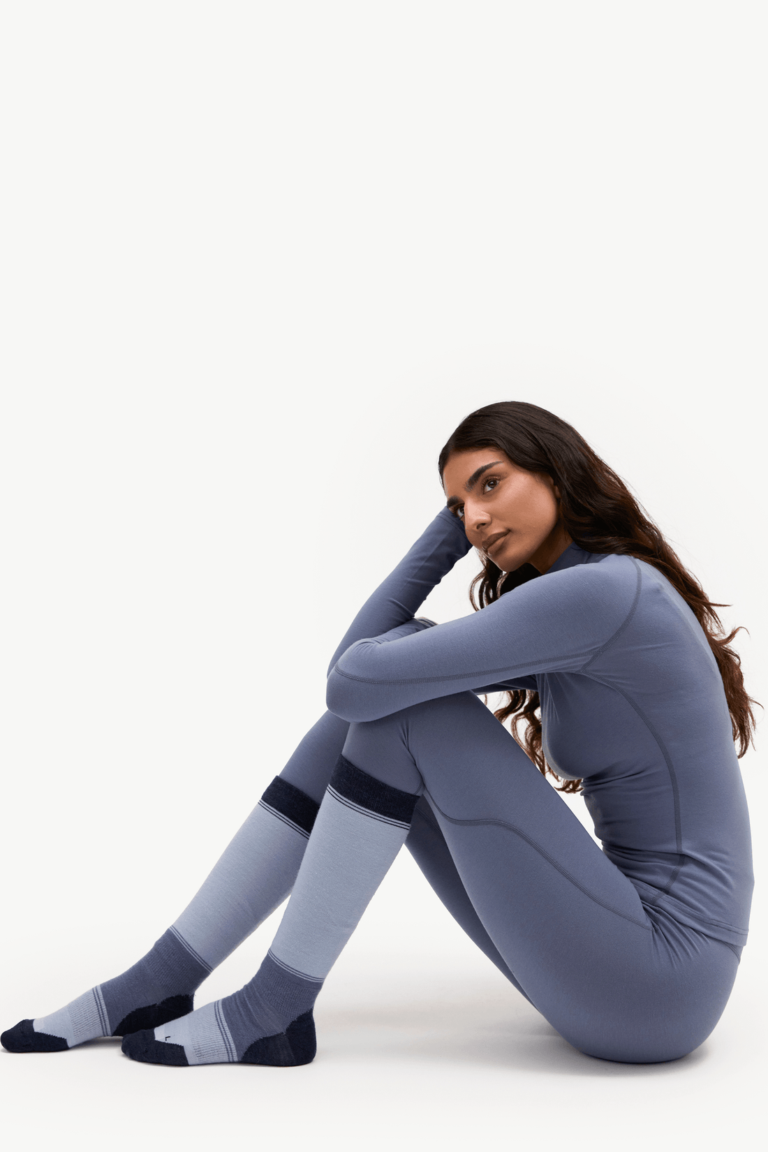 A woman sits on the floor wearing a fitted long-sleeve athletic jumpsuit and matching knee-high socks against a plain white studio background.