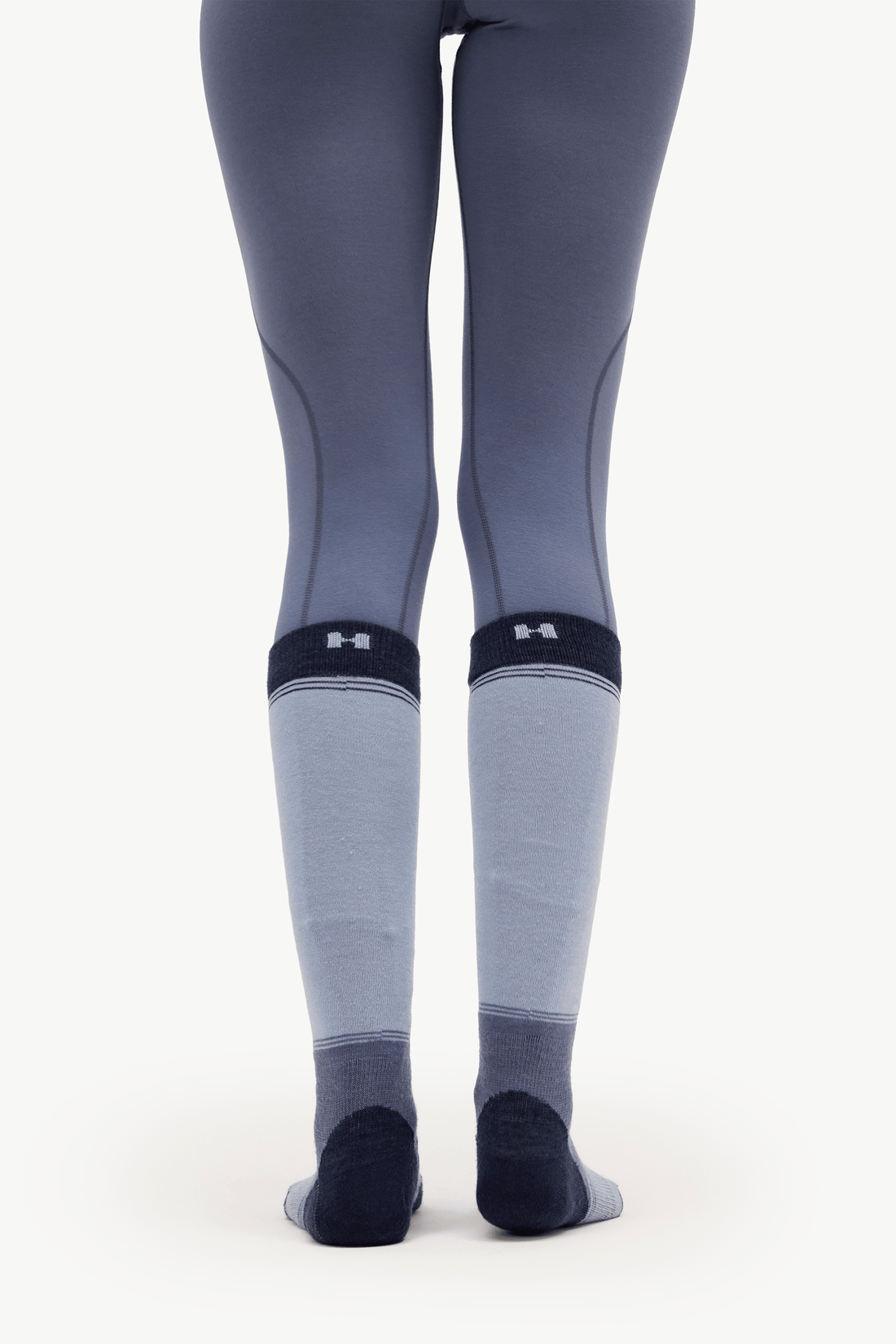 A person shown from the back wearing gray athletic leggings and light gray knee-high compression socks with dark navy heels, toes, and cuffs. Under Armour logos are visible on the socks.